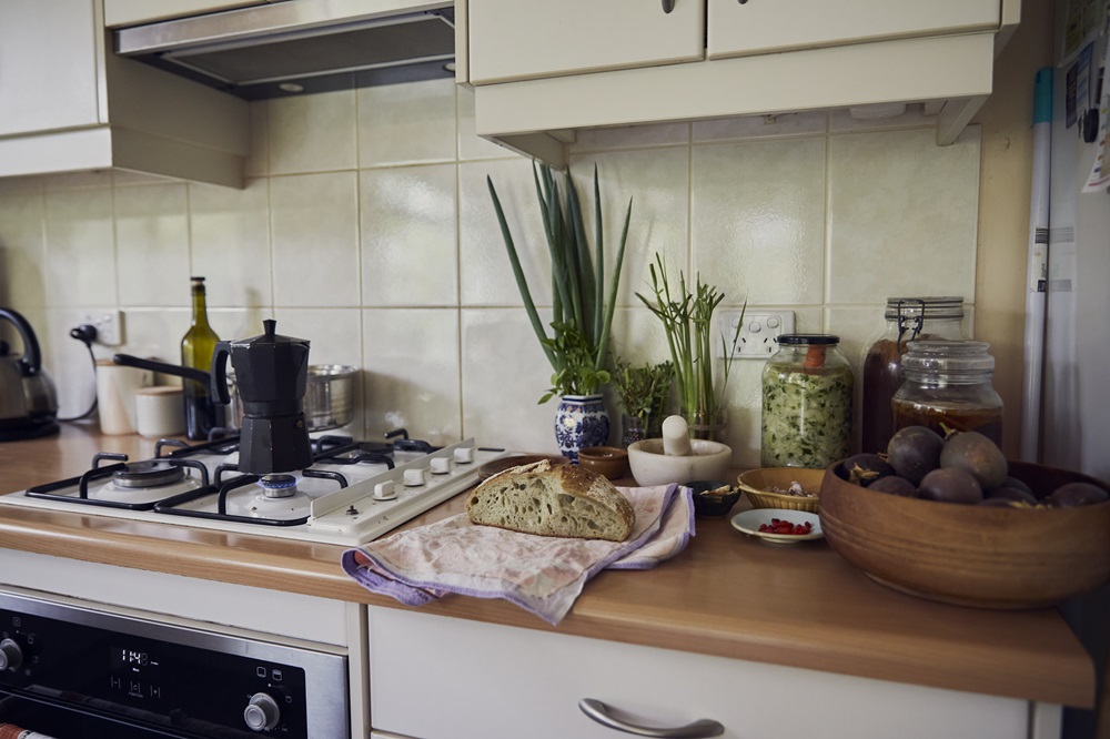 A permaculture kitchen bench scene, showing sourdough, pickles, homegrown figs and veggies, and coffee bubbling on the stovetop. It's the home office of someone who applies permaculture in business.