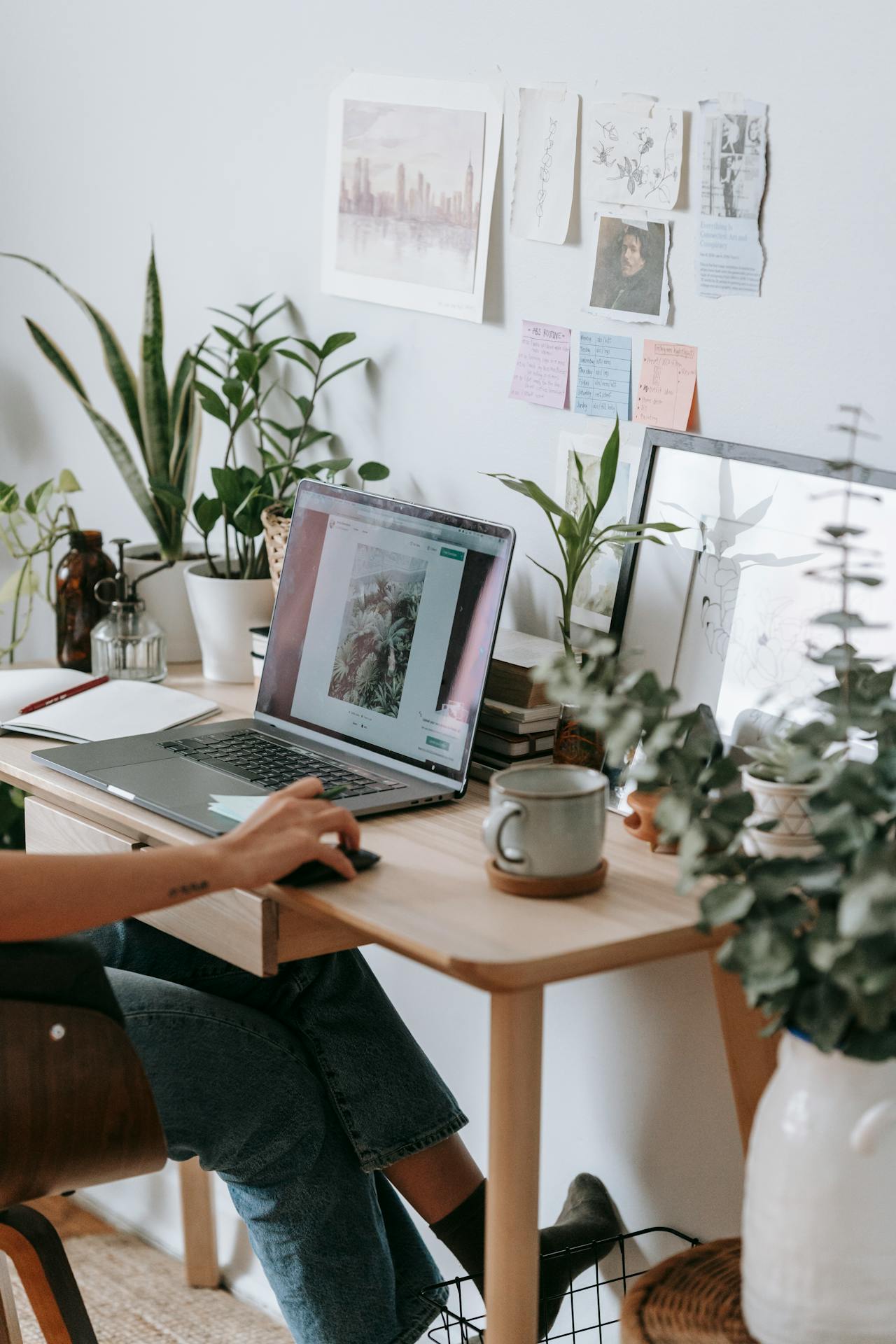 A computer sitting atop a wooden table, surrounded by pot plants, with a woman's hand driving the mouse.