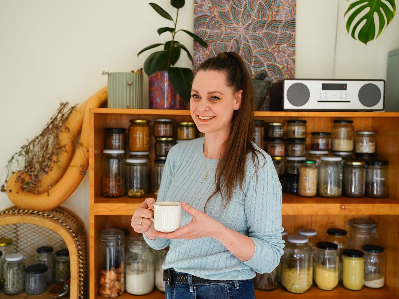 Koren Helbig holding a cup of tea in front of her low-waste pantry, full of jars of bulk dried goods - photo Liza Savchuk